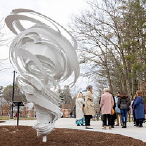 A group of people stands near a white abstract sculpture outside, surrounded by trees on a cloudy day.