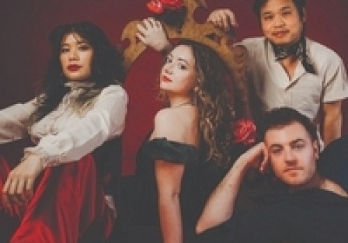 A group of four people in theatrical attire pose confidently against a dark red background.