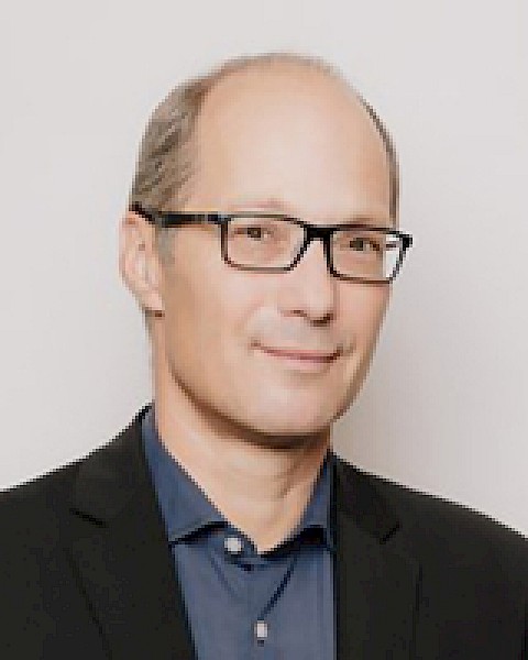 The image shows a person wearing glasses, a dark blazer, and a blue shirt, posing against a plain background.