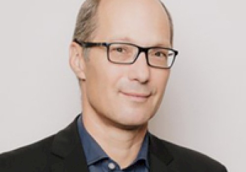 The image shows a person wearing glasses, a dark blazer, and a blue shirt, posing against a plain background.