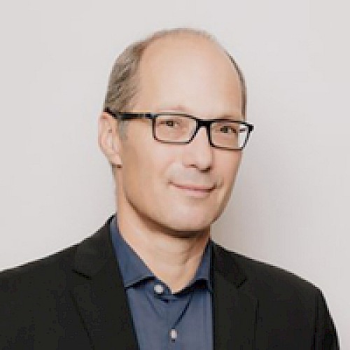 The image shows a person wearing glasses, a dark blazer, and a blue shirt, posing against a plain background.
