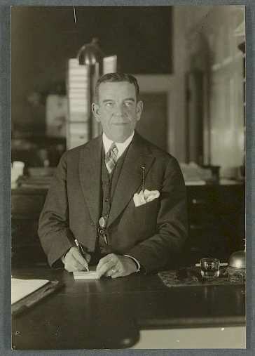 A man in a suit is writing at a desk in an office setting, with various objects around him and a lamp in the background.