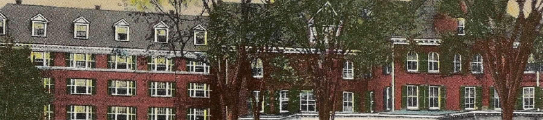 The image shows a large, historic red-brick building with a white porch, surrounded by trees and a grassy area in front.