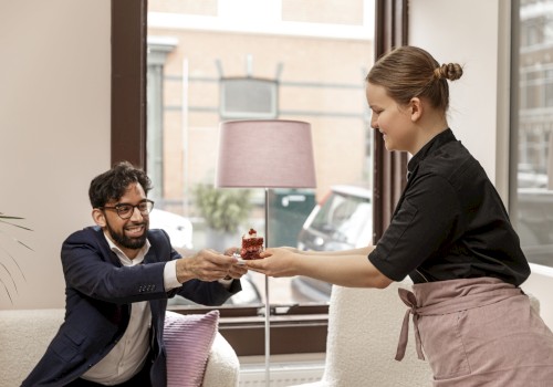 A person is handing another person a small cake in a cozy room with large windows and a lamp in the background.