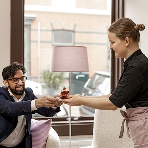 A person is handing another person a small cake in a cozy room with large windows and a lamp in the background.