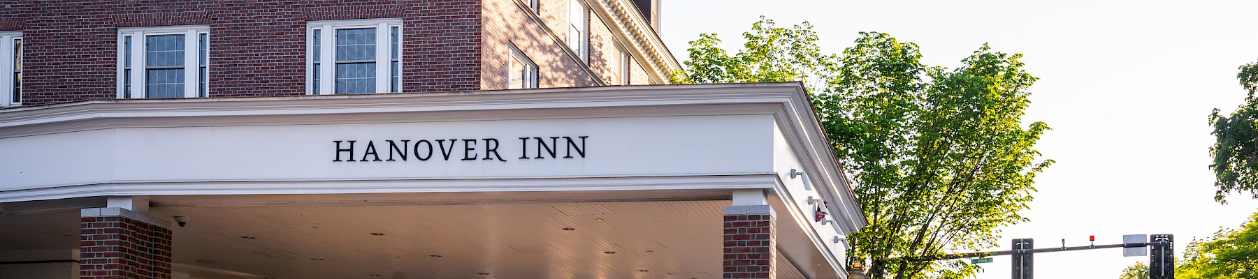 The image shows the entrance of Hanover Inn, a brick building with a covered driveway, trees, and a street with traffic lights nearby.