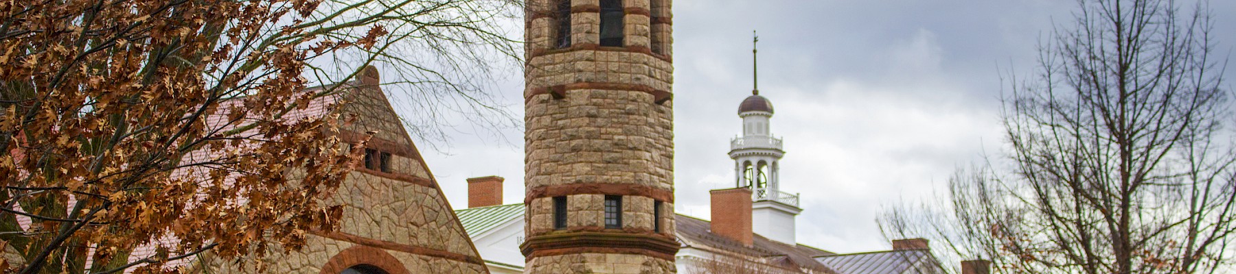 The image shows a historic building with a stone tower, arched windows, and nearby trees with autumn leaves under a cloudy sky.