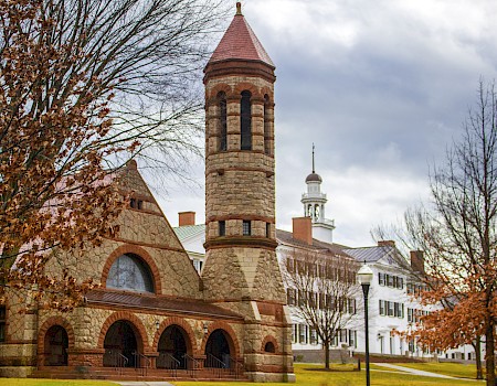 The image shows a historic building with a stone tower, arched windows, and nearby trees with autumn leaves under a cloudy sky.