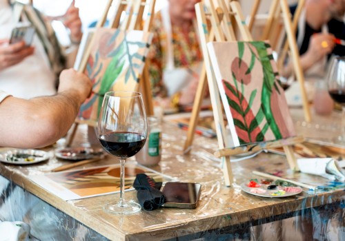 People painting on canvases at easels, with glasses of wine and art supplies on the table in a casual, social setting.