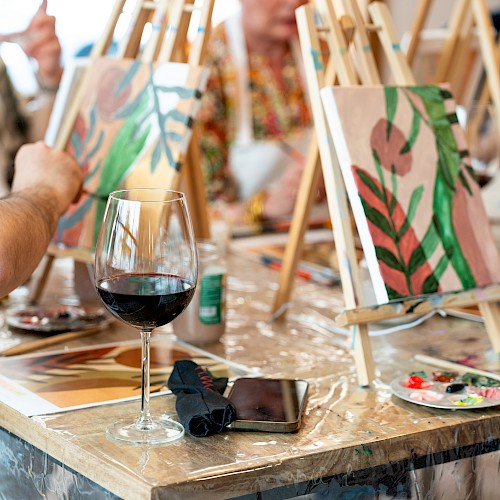People painting on canvases at easels, with glasses of wine and art supplies on the table in a casual, social setting.