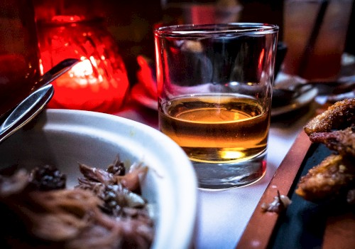 A glass of amber liquid is on a table with food nearby, lit by a red candle creating a cozy ambiance.