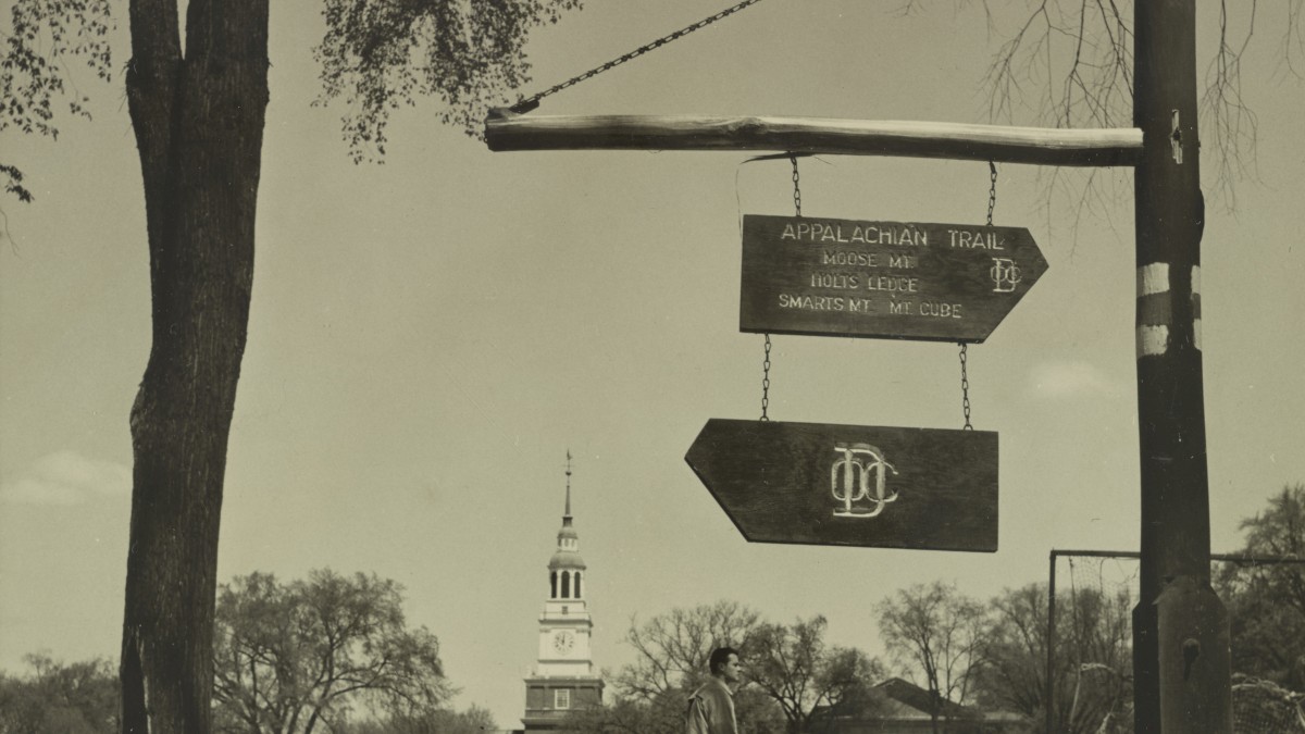 A vintage scene with a directional sign for the Appalachian Trail and a person walking, with college buildings in the background.