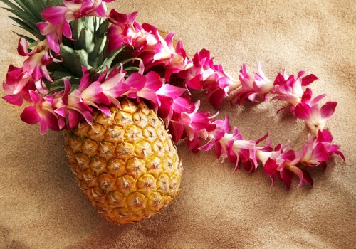 A pineapple with a pink and white flower lei draped over it, resting on a textured beige surface.