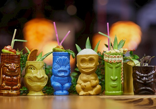 Colorful tiki mugs with straws and garnishes, set against a blurred warm background.