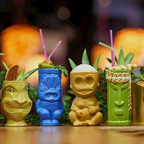 Colorful tiki mugs with straws and garnishes, set against a blurred warm background.