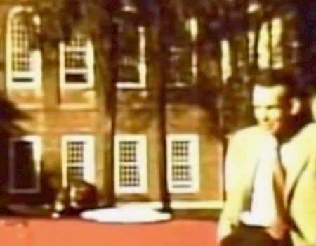Two men in suits stand near a red car in front of a brick building with several windows in the background.