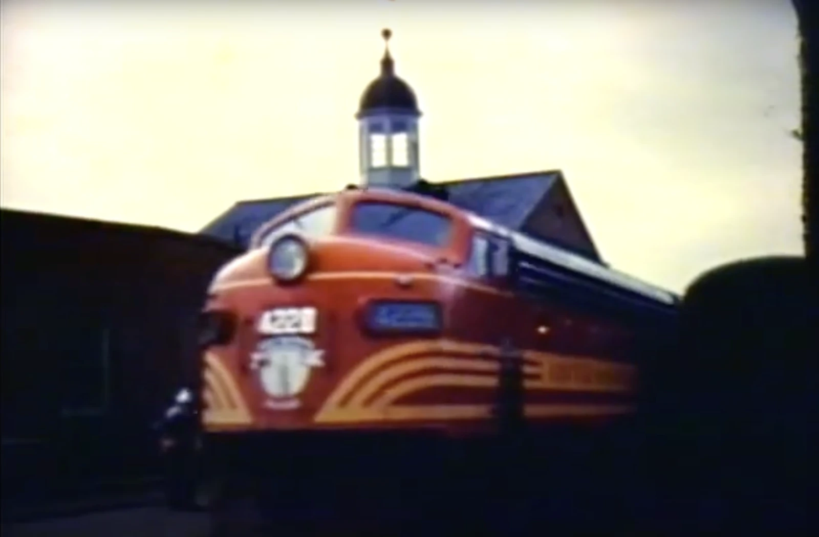 A vintage red and orange locomotive in front of a building with a steeple.