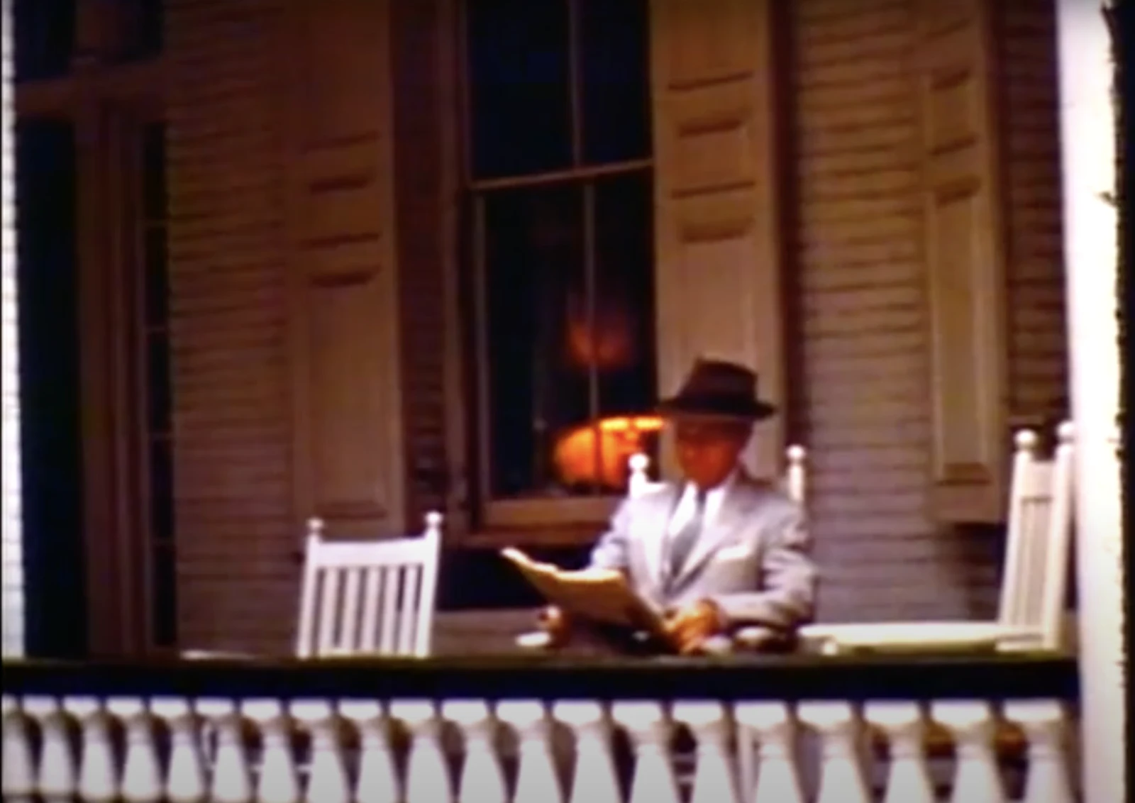 A person in a suit and hat sits on a porch, reading a newspaper, with two white rocking chairs beside them.