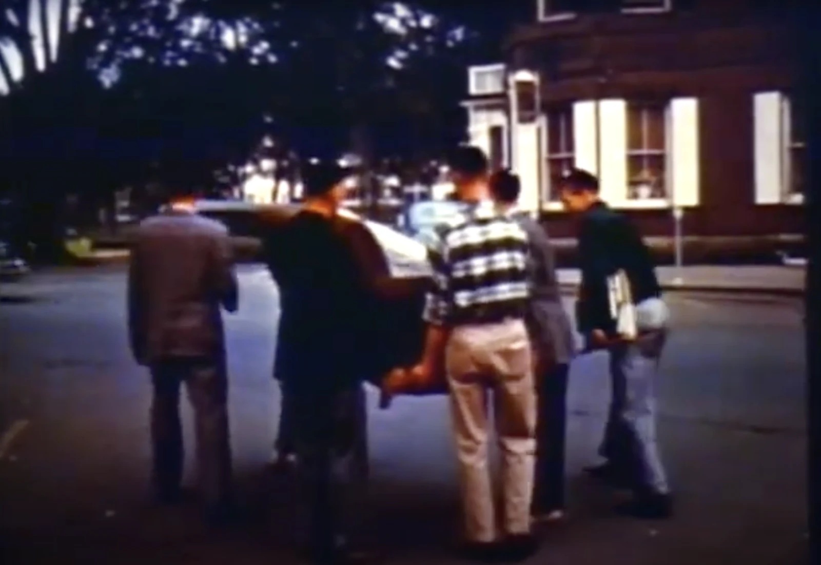 A group of young men are lifting or moving a small car together on a street with trees and buildings in the background.