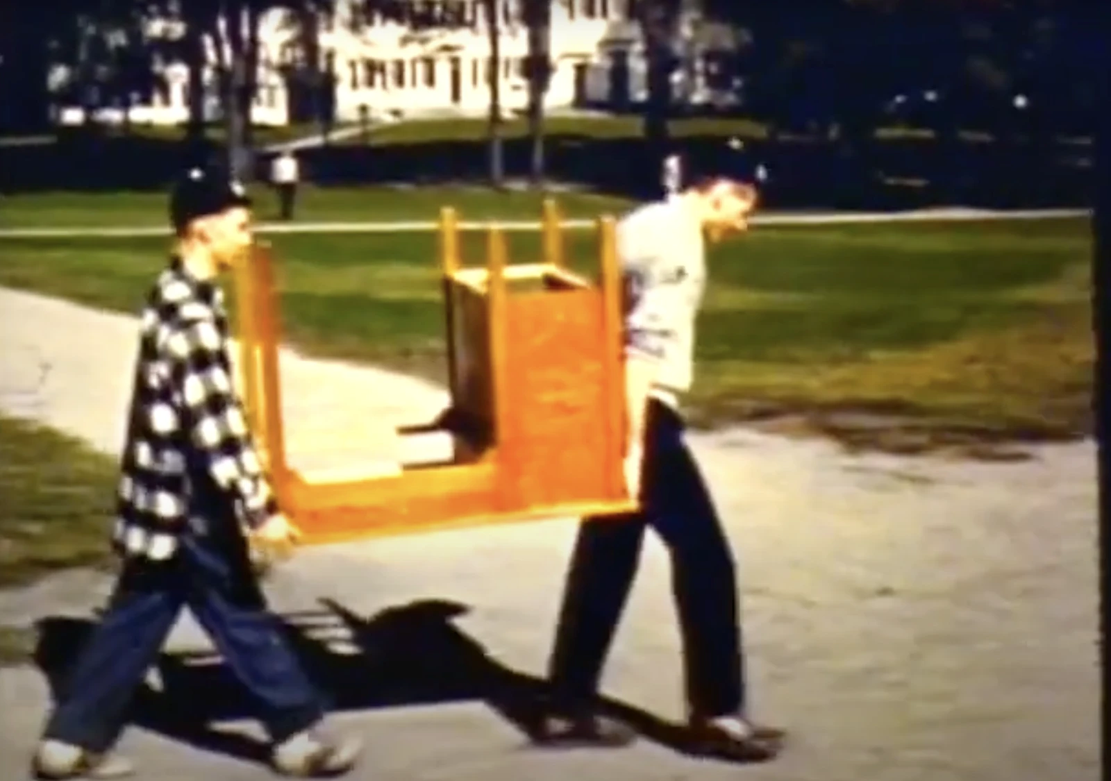 Two people are carrying an upside-down table or chair outdoors, walking on a path under a sunny sky.