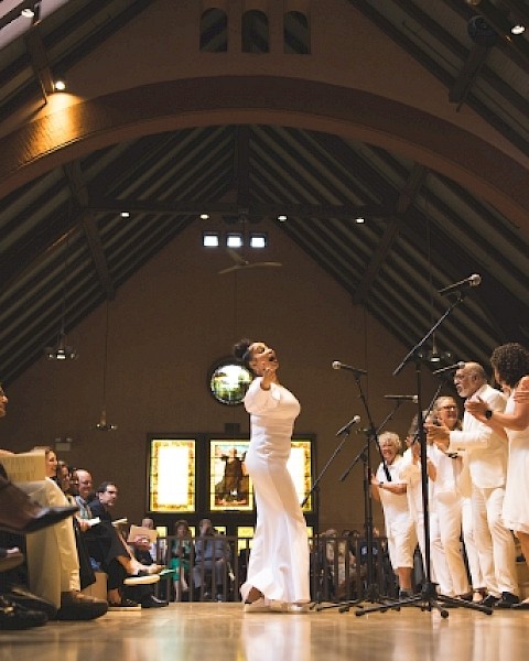 A choir dressed in white is performing energetically on a stage inside a cathedral-like venue, with an audience watching attentively.
