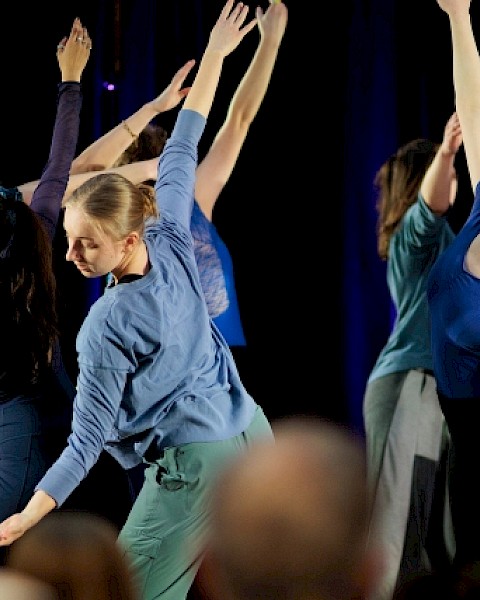 A group of dancers in various poses on stage, wearing blue and dark clothing, perform in front of an audience, arms gracefully extended.