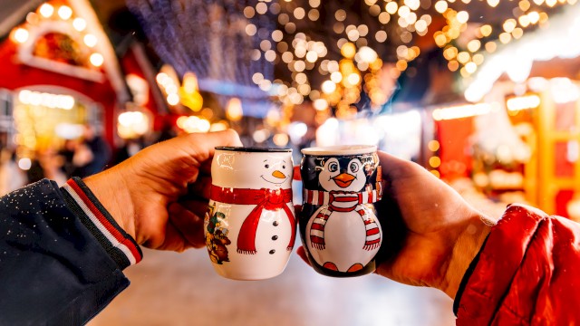 Two hands holding festive mugs with snowman and penguin designs, against a backdrop of warm lights and a winter market scene.