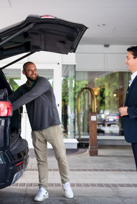 A man in a suit is smiling as another man loads luggage into a car, standing outside a building with brick and glass features.