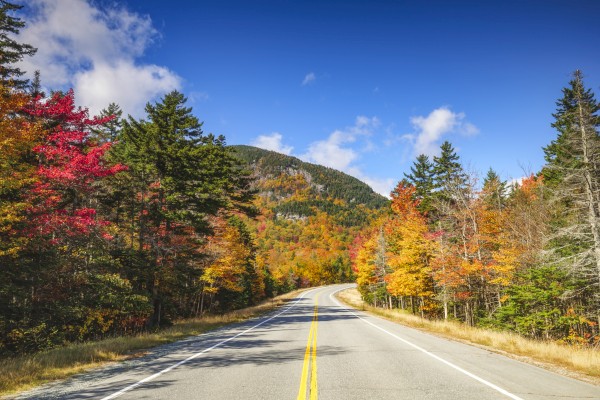 A scenic mountain road winds through colorful autumn foliage on a sunny day with a blue sky and scattered clouds.