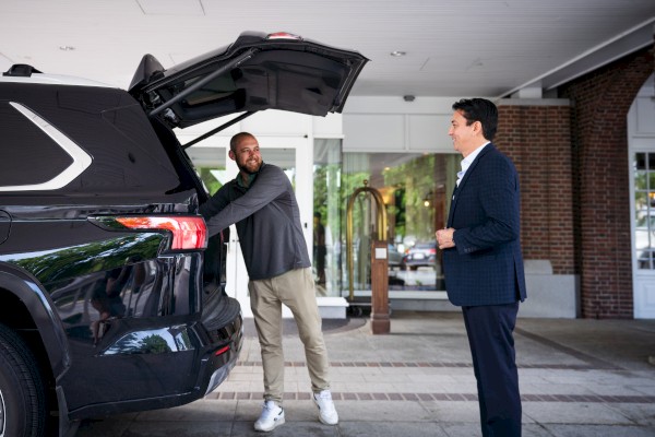 A man is showing a car's trunk to another man in a business setting outside a building.