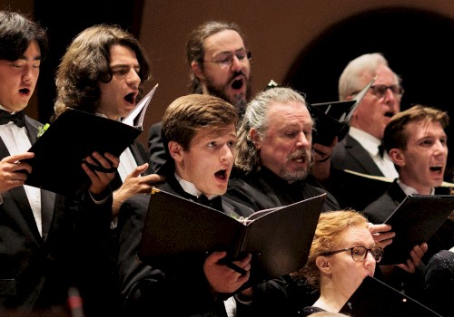 A choir of diverse individuals passionately sings from sheet music during a performance.