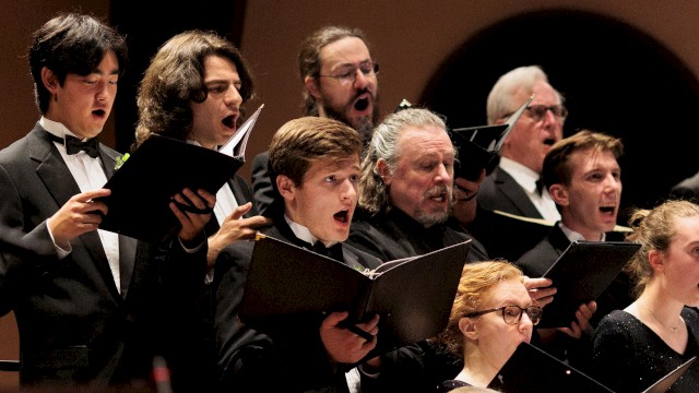 A choir of diverse individuals passionately sings from sheet music during a performance.