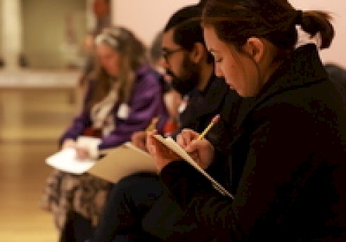 People are seated and writing in notebooks, likely participating in a workshop or class, focusing intently.