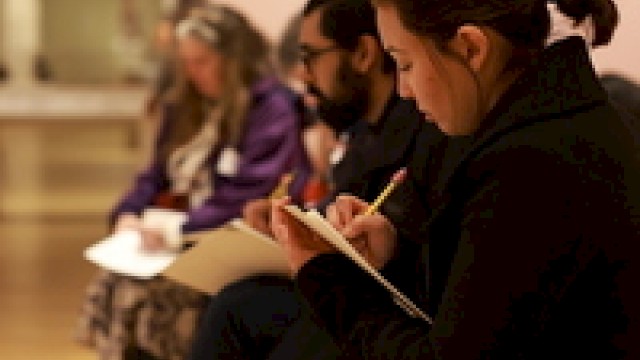 People are seated and writing in notebooks, likely participating in a workshop or class, focusing intently.