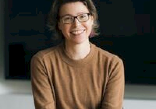 A person wearing glasses and a brown sweater is sitting and smiling, hands clasped.