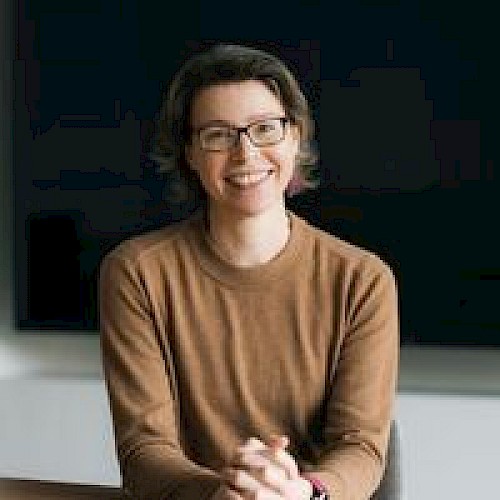 A person wearing glasses and a brown sweater is sitting and smiling, hands clasped.