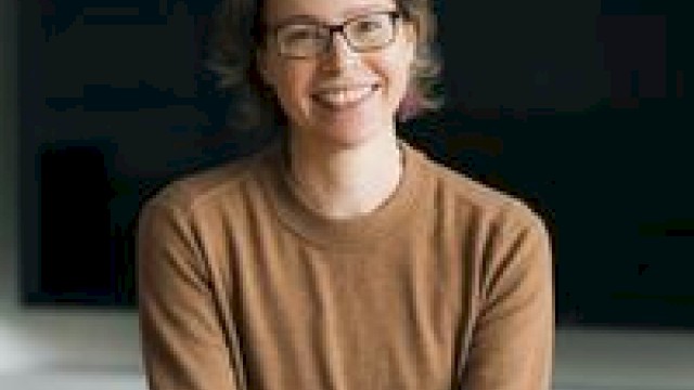 A person wearing glasses and a brown sweater is sitting and smiling, hands clasped.