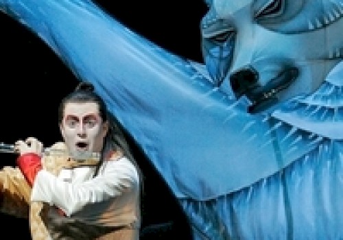 A performer in theatrical costume gestures dramatically alongside a large, stylized blue bear figure on stage.