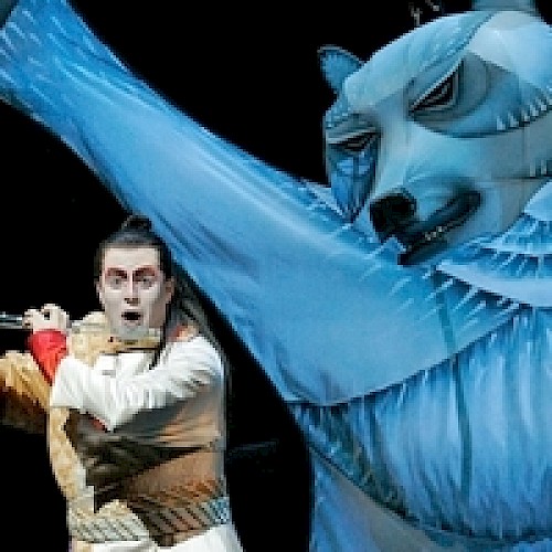 A performer in theatrical costume gestures dramatically alongside a large, stylized blue bear figure on stage.