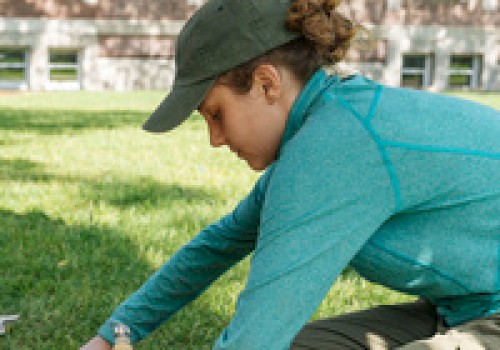 A person in outdoor gear is handling a tool in a grassy area, possibly conducting a task or research.