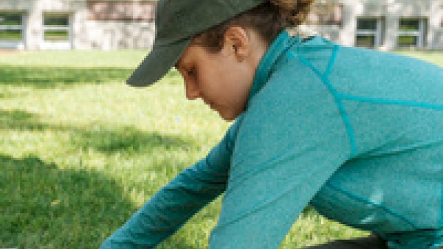 A person in outdoor gear is handling a tool in a grassy area, possibly conducting a task or research.