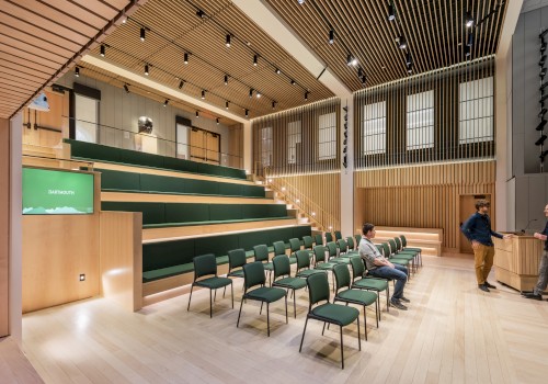A modern auditorium with tiered seating, green chairs, wooden walls, and ceiling, with three people conversing in the space.