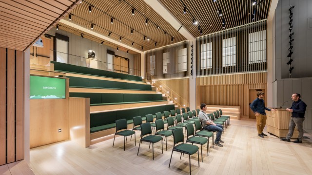 A modern auditorium with tiered seating, green chairs, wooden walls, and ceiling, with three people conversing in the space.