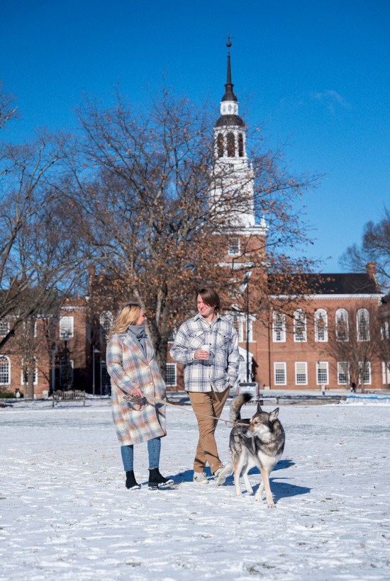A family wearing plaid coats chats with a husky in a snowy yard, a brick campus with a tall steeple rises in the background, bright blue sky.