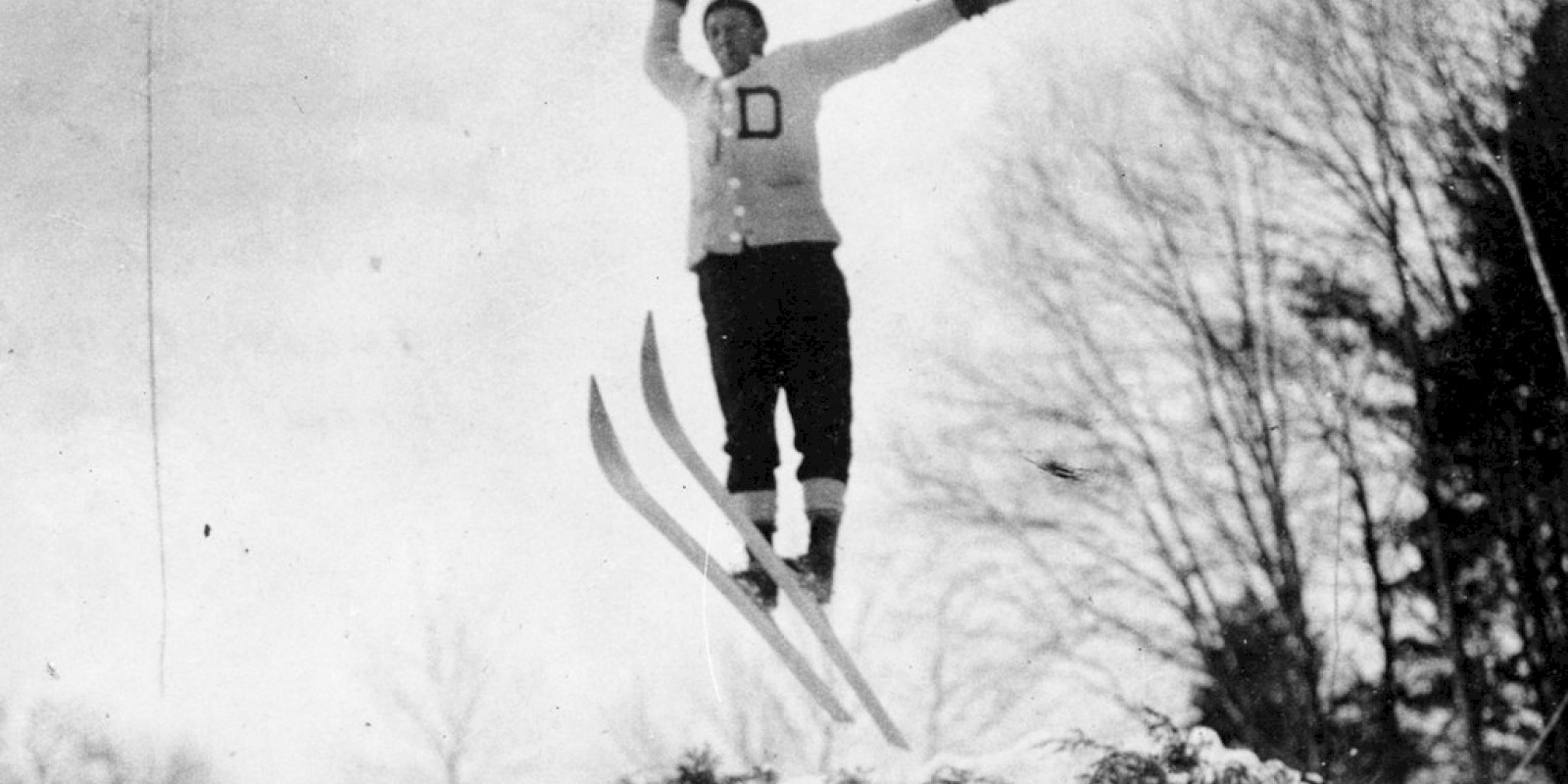 A person in mid-air wearing ski gear skis down a snowy jump, arms raised in triumph, with trees in the background.
