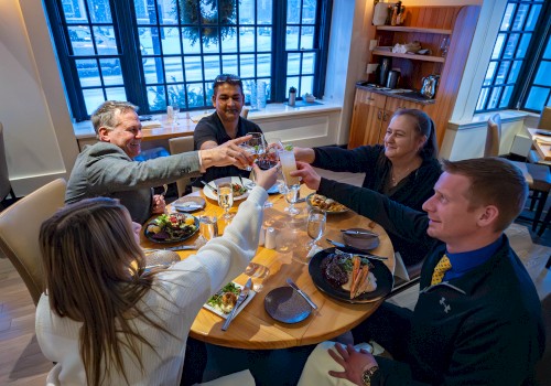 A group of five friends raises glasses in a toast around a round table at a cozy restaurant, enjoying food and conversation.