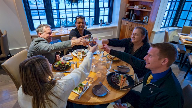 A group of five friends raises glasses in a toast around a round table at a cozy restaurant, enjoying food and conversation.