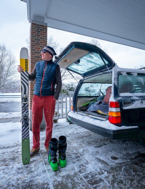 A person dressed for winter stands beside a green ski boot pair near an open car trunk at a snowy gas station or loading area, ready to ski.