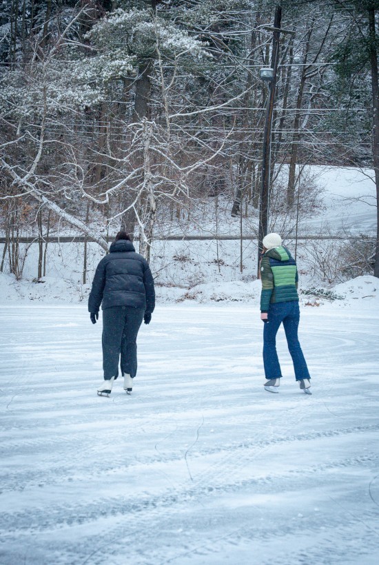 Two people ice-skating on a snowy, wooded area with bare trees and a snow-covered path, enjoying a chilly outdoor winter scene.