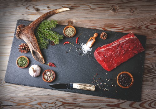 On a slate board: a large raw steak, a deer antler, small bowls with spices, garlic, chili, pine cones, and a knife, all arranged for a rustic meat prep scene.